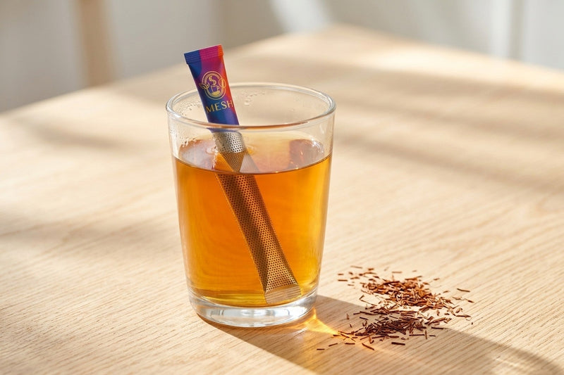 Bir bardak çayda demlenen Mesh Stick çubuğu, yanında rooibos yaprakları